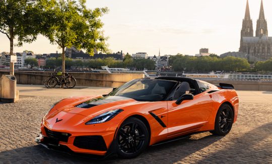 Orange Corvette C7 ZR1 parked on a cobblestone promenade at sunset, with the Cologne Cathedral and the Rhine River in the background