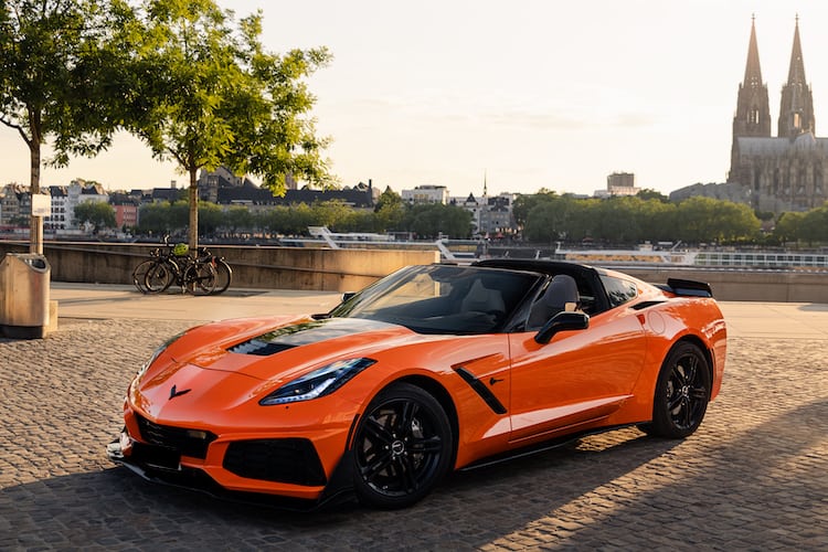 Orange Corvette C7 ZR1 parked on a cobblestone promenade at sunset, with the Cologne Cathedral and the Rhine River in the background