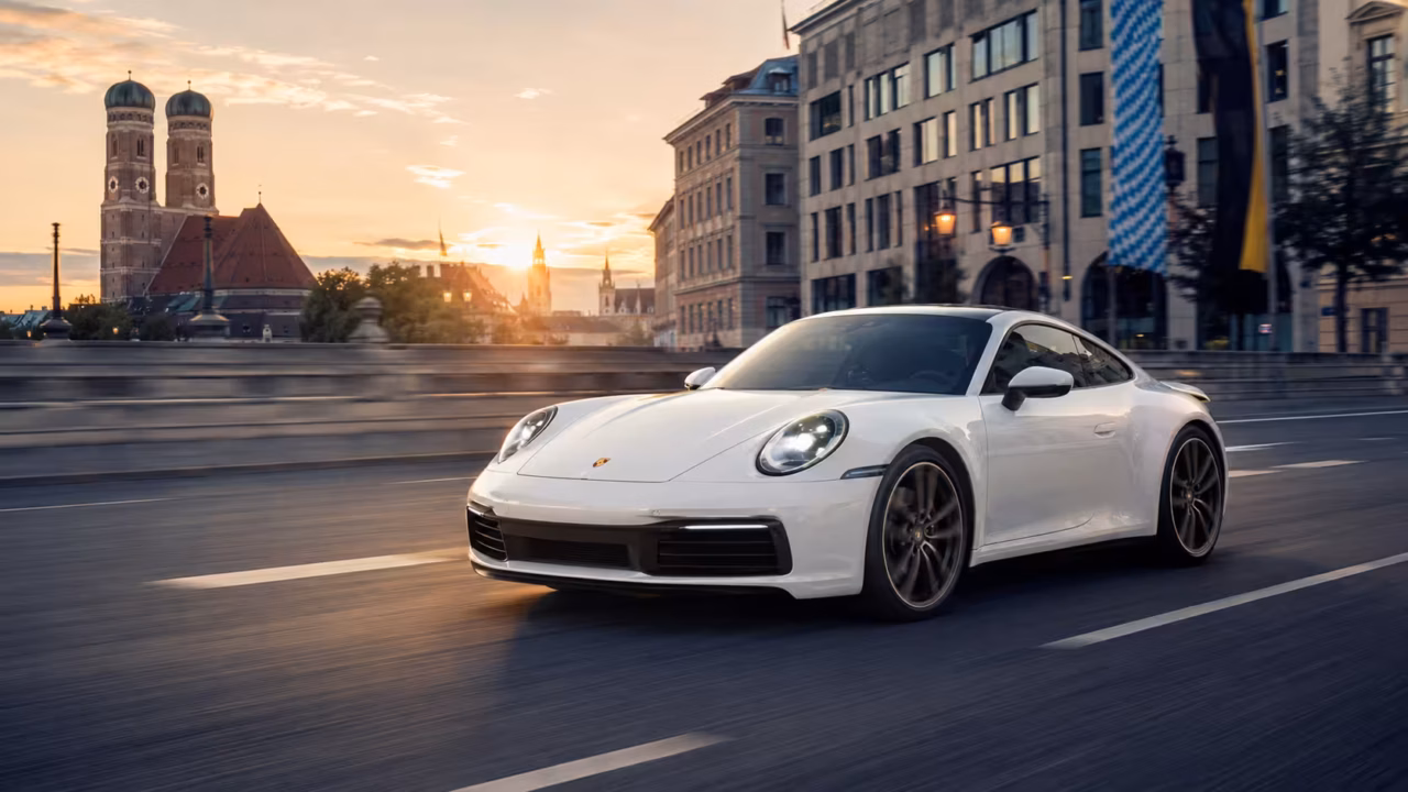 A white Porsche 911 (992 generation) driving across a bridge in Munich during a golden hour sunset, with the historic Frauenkirche towers visible in the background.