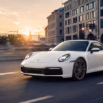 A white Porsche 911 (992 generation) driving across a bridge in Munich during a golden hour sunset, with the historic Frauenkirche towers visible in the background.
