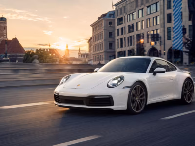A white Porsche 911 (992 generation) driving across a bridge in Munich during a golden hour sunset, with the historic Frauenkirche towers visible in the background.