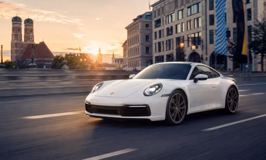 A white Porsche 911 (992 generation) driving across a bridge in Munich during a golden hour sunset, with the historic Frauenkirche towers visible in the background.